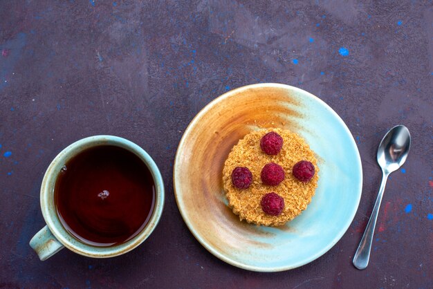 Top view of little round cake with fresh raspberries inside plate with tea on the dark-blue surface