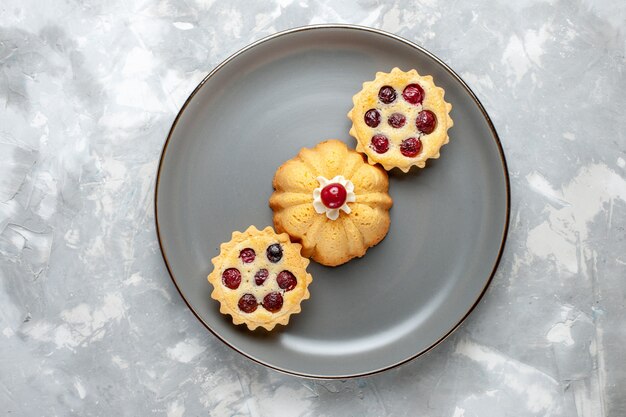 Top view little yummy cakes with sugar powder and cherries on the light desk fruit cake biscuit sweet color