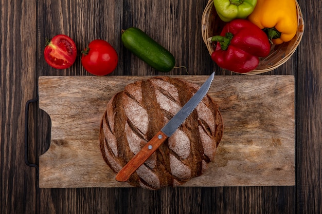 Free photo top view loaf of black bread on a stand with a knife  with tomatoes  cucumber and bell peppers on a wooden background