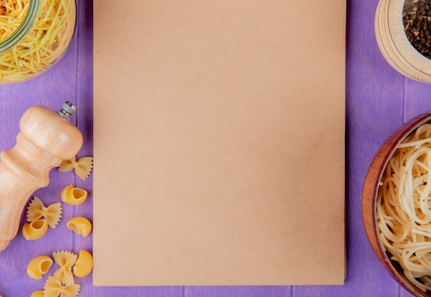 Free photo top view of macaronis as cooked and uncooked spaghetti farfalle pipe-rigate with black pepper around note pad on purple background with copy space