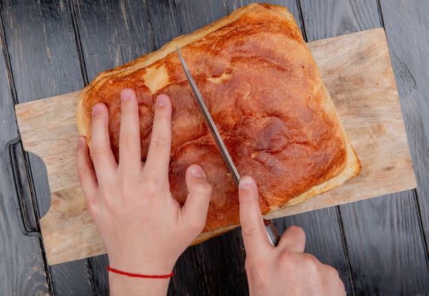 top view of male hands cutting homemade bread with knife on cutting board on wooden background