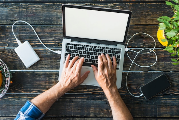 Top view of man's hand typing on laptop connected with power bank and cellphone