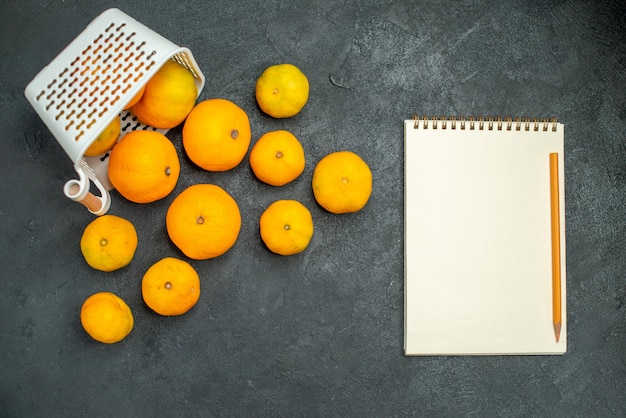 Free photo top view mandarines and oranges scattered from plastc basket notebook pencil on dark surface