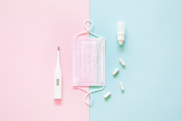 Top view of medical mask, pills and thermometer on pink and green background.