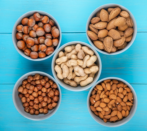 Top view of mixed nuts in shell and without shell in bowls almond hazelnuts and peanuts on blue background