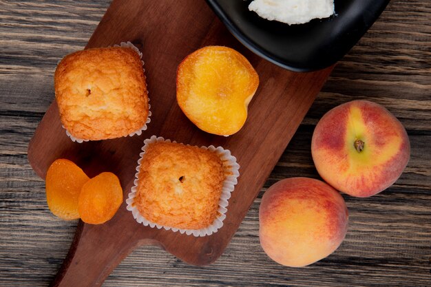 Top view of muffins with dried apricots on wood cutting board and fresh peaches on rustic