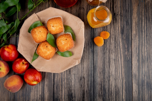 Top view of muffins with green leaves on craft brown paper with fresh ripe nectarines and peach jam in a glass jar on rustic wood with copy space