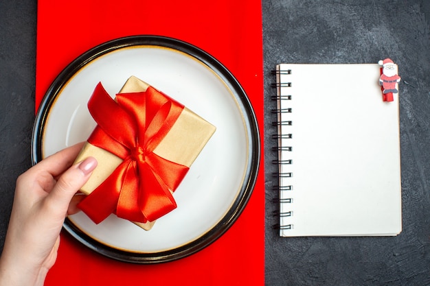 Free photo top view of national christmal meal background with hand holding empty plates with bow-shaped red ribbon on a red napkin and notebook on black table