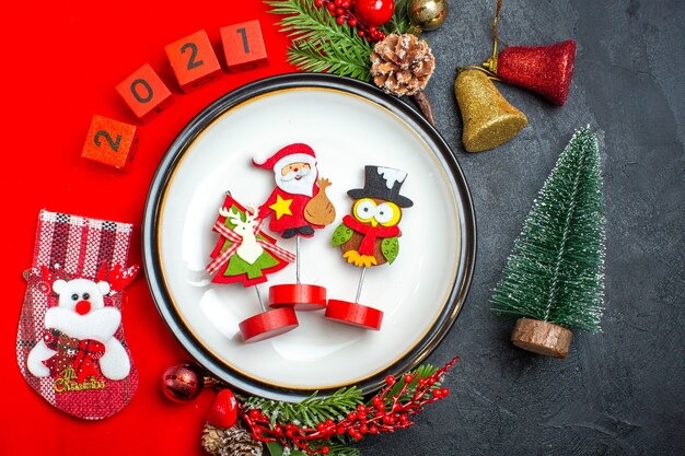 Top view of New year background with dinner plate decoration accessories fir branches and numbers christmas sock on a red napkin next to christmas tree on a black table