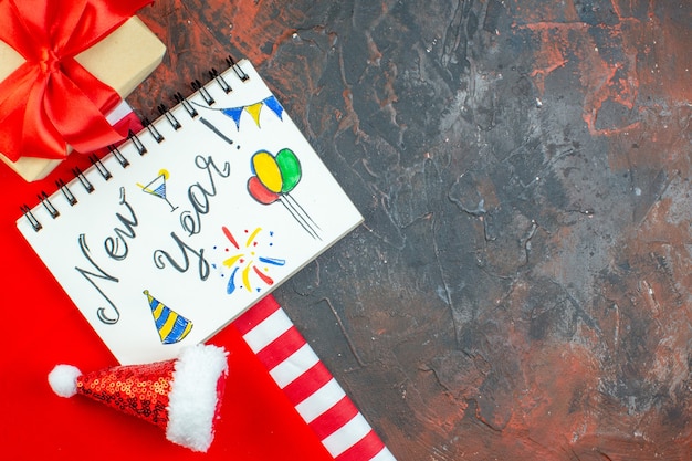 Top view new year written on notepad mini santa hat on red tablecloth on dark red table free space