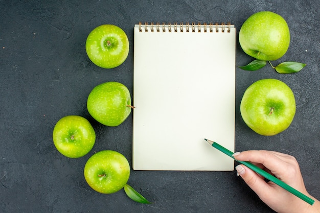 Top view notebook green apples green pencil in female hand on dark surface