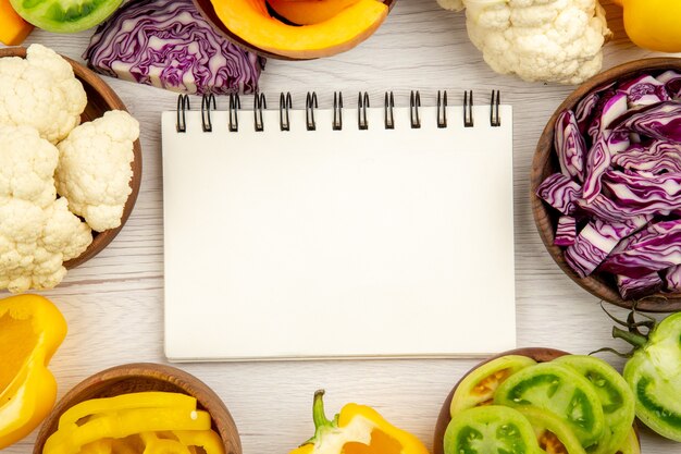 Top view notebook red cabbage in bowl cauliflower yellow bell pepper green tomato on white wooden surface