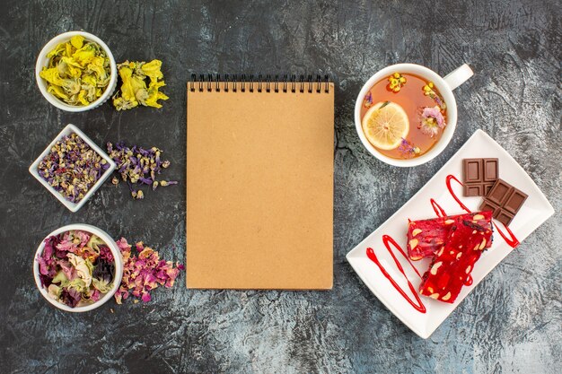 Top view of notebook with bowls of dry flowers and a cup of tea near a plate of chocolate