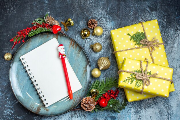 Top view of notebook with pen blue plate decorative accessories conifer cone next to yellow gift boxes on dark background