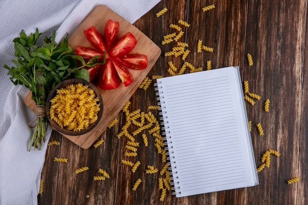 Free photo top view of notebook with raw pasta in a bowl with slicing tomato slices on a cutting board with a bunch of mint on a wooden surface