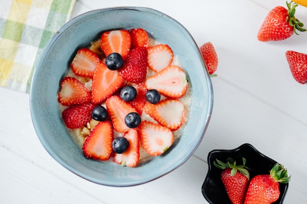 Top view of oatmeal porridge with sliced strawberries and blueberries in a ceramic bowl and overnight oats in a glass jar on the table