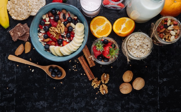 Top view of oatmeal porridge with strawberries blueberries bananas dried fruits and nuts in a ceramic bowl and glass jars with mixed nuts overnights oats and oat flakes on the table