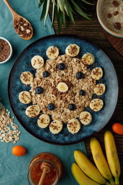 Top view of oatmeal with banana blackthorn walnut and sesame in plate with jam oat kumquat pineapple leaves on blue cloth on wooden background