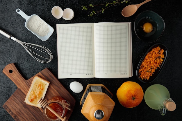 Top view of open notepad with flour egg orange lime juice whisk spoon jam bread slice orange zest grater on black background