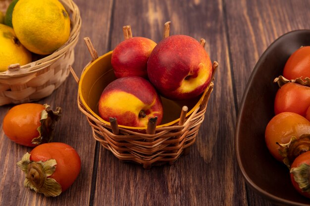 Top view of organic peaches on a bucket with tangerines with persimmons isolated on a wooden wall