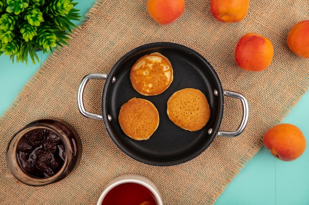 Top view of pancakes in pan and strawberry jam tea apricots on sackcloth on blue background