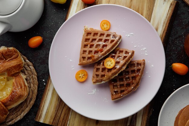 Top view of pancakes with kumquat slices in plate on cutting board with croissant whole kumquats and teapot on black background