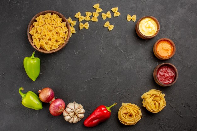 Top view pasta pasta in bowl colorful sauces green and red ball pepper garlic onion are laid out in a circle on the black table