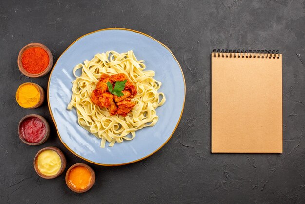 Free photo top view pasta and sauces bowls of five kind of colorful sauces and pasta with gravy herbs and meat next to the cream notebook on the dark table