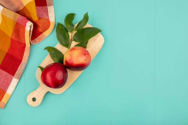 Top view of peaches and leaves on cutting board with plaid cloth on blue background with copy space