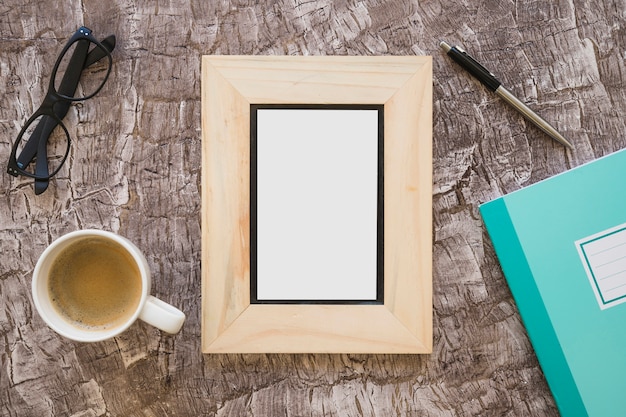 Top view of picture frame; coffee cup; eyeglasses; pen and notebook on textured background