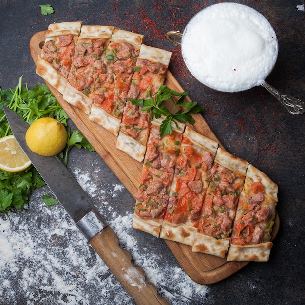 Top view pide with pieces of meat and parsley and lemon and knife and ayran in cutting board