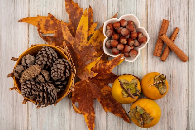 Top view of pine cones on a bucket with leaves with hazelnut on a bowl with cinnamon sticks on a grey wooden table