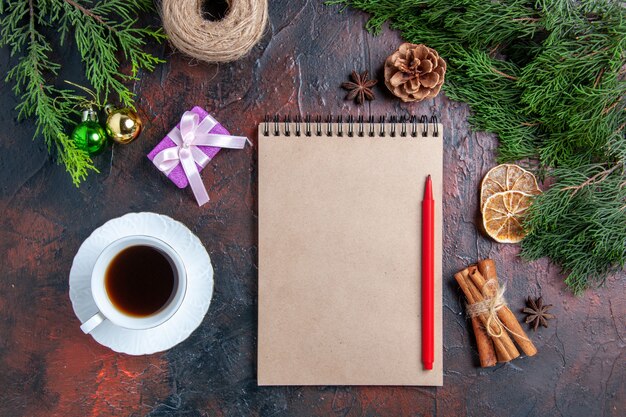 Top view pine tree branches and pinecones a notebook red pen anises cinnamon cup of tea on dark red surface