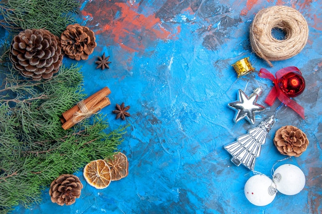 Top view pine tree branches with cones cinnamon sticks anise seeds dried lemon slices and vertical row straw thread xmas tree toys on blue-red surface