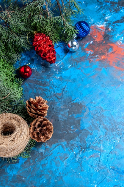 Top view pine tree branches with pinecones and colorful xmas tree toys straw thread on blue-red surface