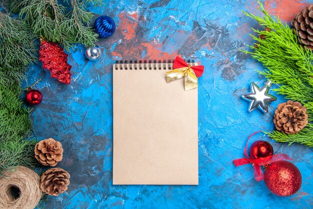 Top view pine tree branches with pinecones and colorful xmas tree toys straw thread a notebook on blue-red surface