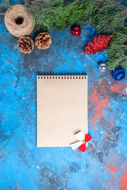 Top view pine tree branches with pinecones and colorful xmas tree toys straw thread notebook with bow on blue-red background