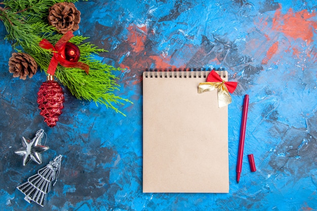 Top view pine tree branches with pinecones hanging ornaments notebook and red pen on blue-red surface