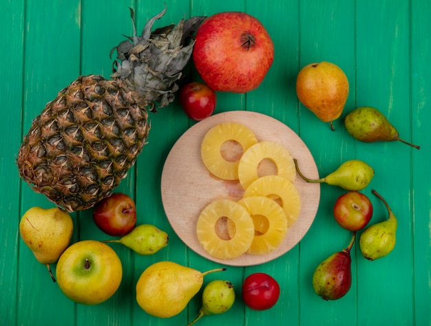 Top view of pineapple slices on cutting board and pineapple pomegranate peach plum apple on green surface