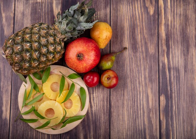 Top view of pineapple slices with leaves on cutting board and pineapple pomegranate peach plum on wooden surface