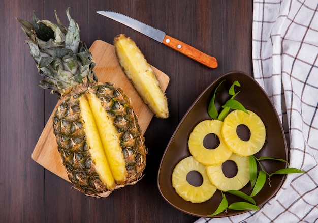 Top view of pineapple with one piece cut out from whole fruit on cutting board with pineapple slices and knife on wooden surface