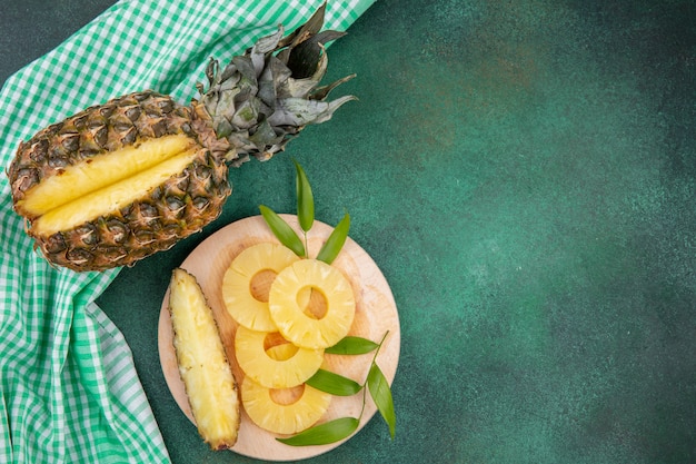 Free Photo top view of pineapple with one piece cut out from whole fruit and pineapple slices on cutting board on plaid cloth and green surface