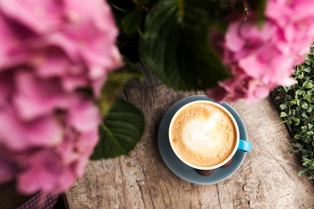 Top view of pink flower and tasty coffee with frothy foam on wooden surface