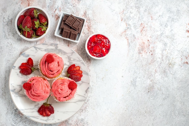 Top view of pink strawberry cakes with jam and chocolate bars on a white surface