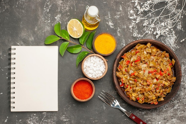 Free photo top view plate of beans and spices bowls of colorful spices lemon white notebook the plate of green beans next to the bottle of oil and fork on the dark table