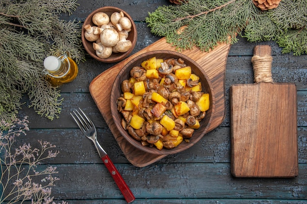 Top view plate and cutting board potatoes and mushrooms in bowl on brown board next to the fork and wooden cutting board under bowl of mushrooms oil in bottle and branches with cones