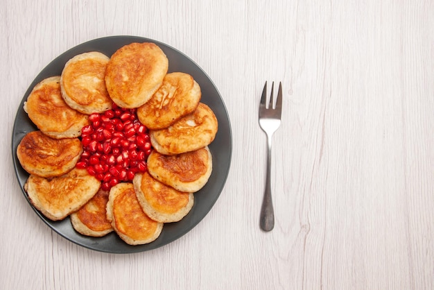 Top view plate of dessert seeds of red pomegranate and pancakes on the black plate next to the fork on the white background