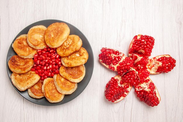 Top view plate of dessert seeds of red pomegranate and pancakes on the black plate next to the peeled pomegranate on the white surface