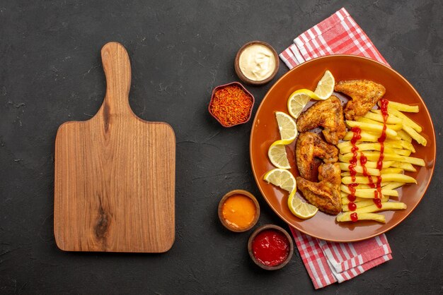 Top view plate of fastfood chicken wings french fries with lemon and ketchup and bowls of sauces and spices on pink-white checkered tablecloth next to the wooden cutting board