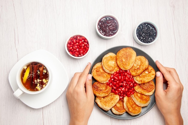 Top view plate in hands white cup of tea with cinnamon sticks bowls of jam and plate of pancakes and pomegranate in hands on the table